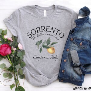May include: Athletic heather gray t-shirt with the text "SORRENTO The Italian Lemon Town Campania, Italy". The shirt features a watercolor illustration of a lemon and leaves. A denim jacket is draped over the shirt.