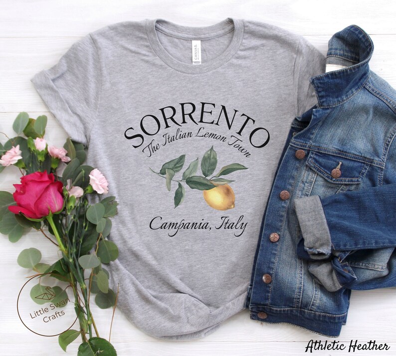 May include: Athletic heather gray t-shirt with the text "SORRENTO The Italian Lemon Town Campania, Italy". The shirt features a watercolor illustration of a lemon and leaves. A denim jacket is draped over the shirt.