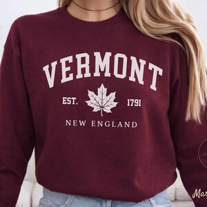 Könnte beinhalten: Burgunderfarbenes Sweatshirt mit dem Wort "VERMONT" in weißen Blockbuchstaben. Darunter ein weißes Ahornblatt und der Text "EST. 1791 NEW ENGLAND". Das Sweatshirt hat einen Rundhalsausschnitt.