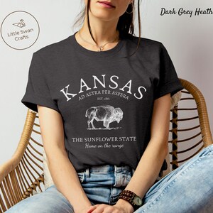 May include: A dark grey heather t-shirt with a white graphic design featuring the text "KANSAS AD ASTRA PER ASPERA EST. 1861" and an illustration of a buffalo. The text "THE SUNFLOWER STATE Home on the range" is also included in the design.