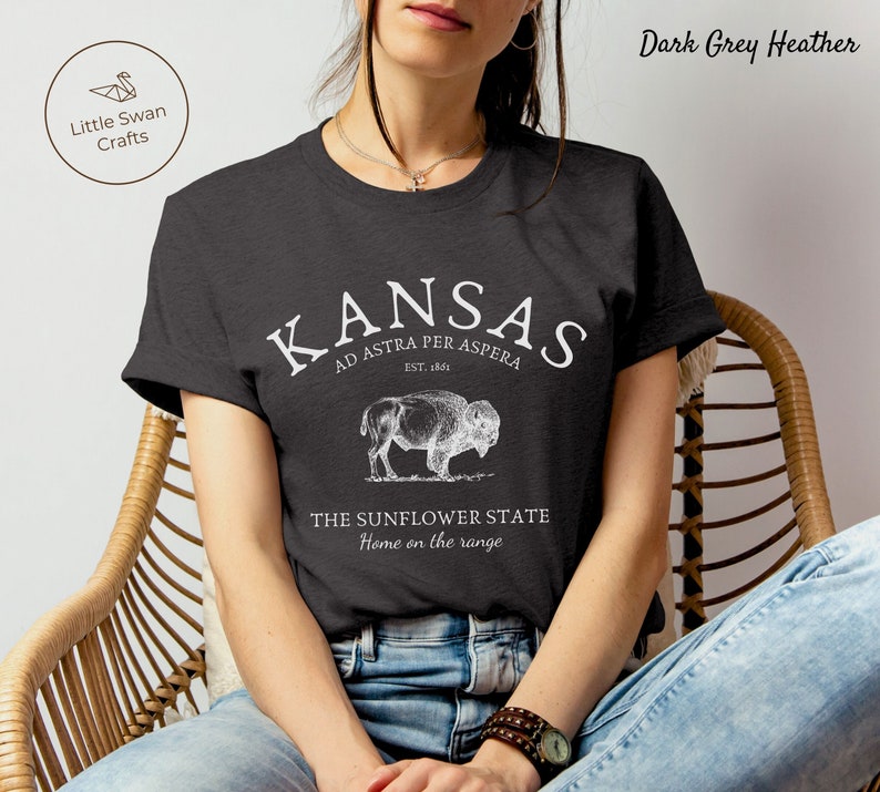 May include: A dark grey heather t-shirt with a white graphic design featuring the text "KANSAS AD ASTRA PER ASPERA EST. 1861" and an illustration of a buffalo. The text "THE SUNFLOWER STATE Home on the range" is also included in the design.