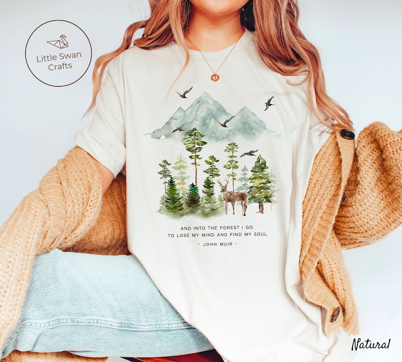Pode incluir: Camiseta branca com uma ilustra&ccedil;&atilde;o em aquarela de uma cena de floresta com montanhas, &aacute;rvores e um veado. O texto "AND INTO THE FOREST I GO. TO LOSE MY MIND AND FIND MY SOUL - JOHN MUIR -" est&aacute; impresso abaixo da ilustra&ccedil;&atilde;o.
