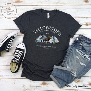 May include: Dark grey heather t-shirt with a Yellowstone National Park graphic featuring mountains, a moose, and the text "Wyoming, Montana, Idaho, Est. 1872." The shirt is paired with blue jeans and black sneakers.