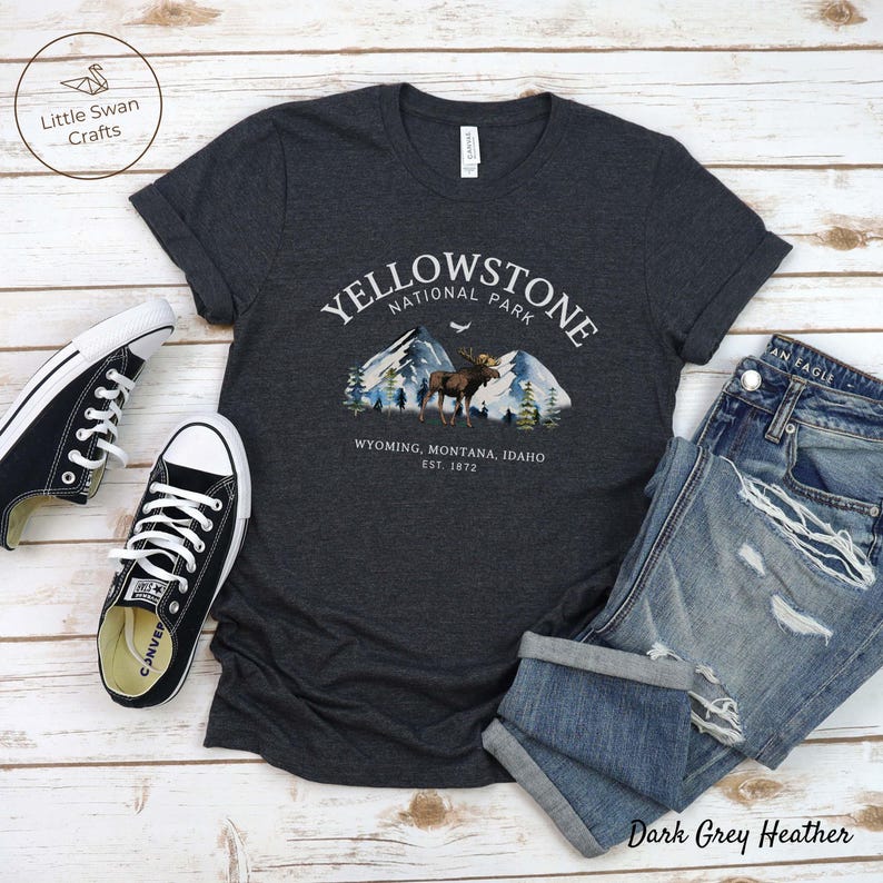 May include: Dark grey heather t-shirt with a Yellowstone National Park graphic featuring mountains, a moose, and the text "Wyoming, Montana, Idaho, Est. 1872." The shirt is paired with blue jeans and black sneakers.