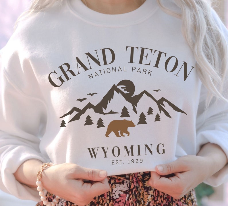 May include: White sweatshirt with a brown graphic of a bear and mountains. The text "Grand Teton National Park" and "Wyoming Est. 1929" is printed on the sweatshirt.