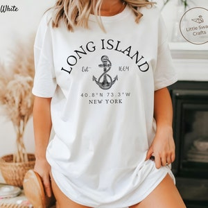 Op de afbeelding: Wit T-shirt met een zwart grafisch ontwerp met de tekst "LONG ISLAND" in een boog boven een anker met de tekst "Est 1664" en "40.8&deg;N 73.3&deg;W NEW YORK" onder het anker.