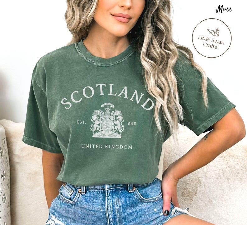 May include: A moss green t-shirt with the word "SCOTLAND" arched across the top. Below the word "SCOTLAND" is a crest with the words "EST. 843" and "UNITED KINGDOM". The shirt is a comfortable, casual style.