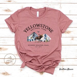 May include: Heather mauve t-shirt featuring a graphic of Yellowstone National Park. The design includes mountains, trees, and a moose, with the text "YELLOWSTONE NATIONAL PARK" and "WYOMING, MONTANA, IDAHO EST. 1872".