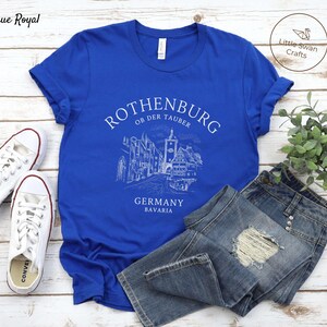 Pode incluir: Uma camiseta azul royal com um design gr&aacute;fico branco da silhueta da cidade de Rothenburg ob der Tauber. O gr&aacute;fico inclui o texto "Rothenburg ob der Tauber", "Alemanha" e "Baviera".