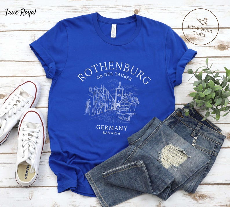 Pode incluir: Uma camiseta azul royal com um design gr&aacute;fico branco da silhueta da cidade de Rothenburg ob der Tauber. O gr&aacute;fico inclui o texto "Rothenburg ob der Tauber", "Alemanha" e "Baviera".