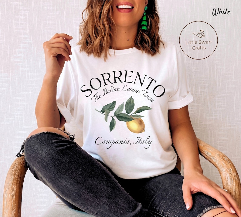 May include: White t-shirt with the text "Sorrento The Italian Lemon Town Campania, Italy" in black script. A watercolor illustration of a lemon branch with green leaves and a yellow lemon is below the text. The shirt has rolled-up sleeves.