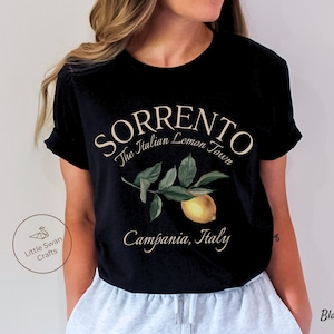 May include: Black t-shirt featuring the text "SORRENTO The Italian Lemon Town" above a watercolor illustration of a lemon and leaves. The text "Campania, Italy" is below the illustration. The shirt also has the text "Black" on the bottom right.