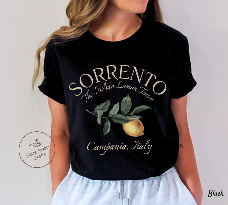 May include: Black t-shirt featuring the text "SORRENTO The Italian Lemon Town" above a watercolor illustration of a lemon and leaves. The text "Campania, Italy" is below the illustration. The shirt also has the text "Black" on the bottom right.