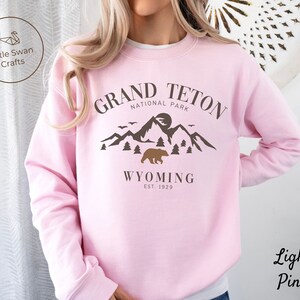 May include: Light pink sweatshirt with a brown graphic design of mountains, a bear, and the text "Grand Teton National Park Wyoming Est. 1929".