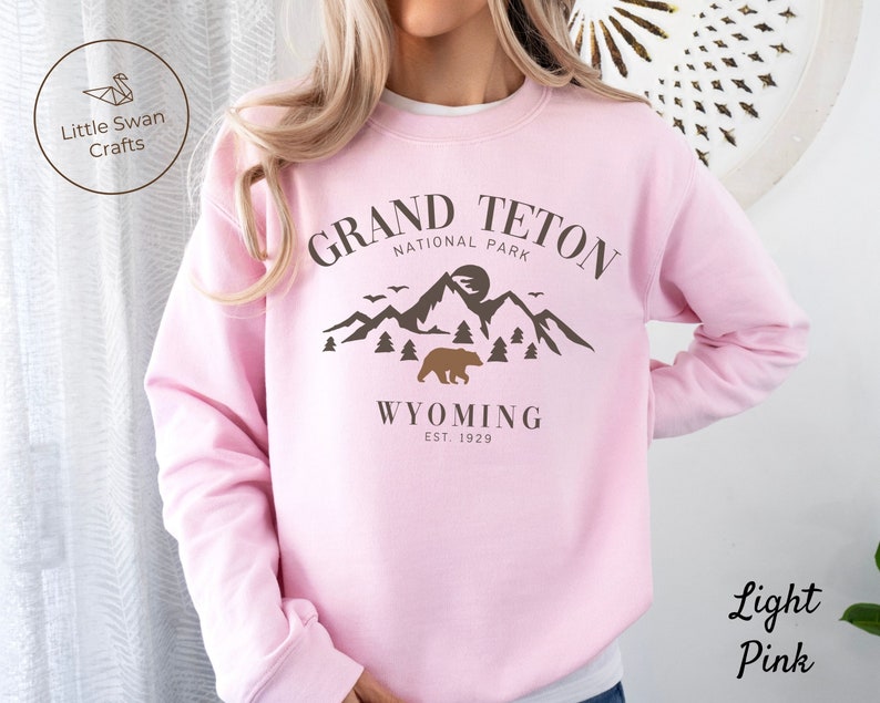 May include: Light pink sweatshirt with a brown graphic design of mountains, a bear, and the text "Grand Teton National Park Wyoming Est. 1929".