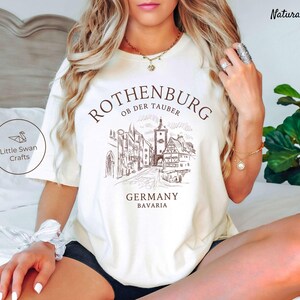 Pode incluir: Camiseta branca com um gr&aacute;fico marrom da cidade de Rothenburg ob der Tauber na Alemanha. O texto "Rothenburg ob der Tauber" est&aacute; acima do gr&aacute;fico, e o texto "Alemanha Baviera" est&aacute; abaixo do gr&aacute;fico.