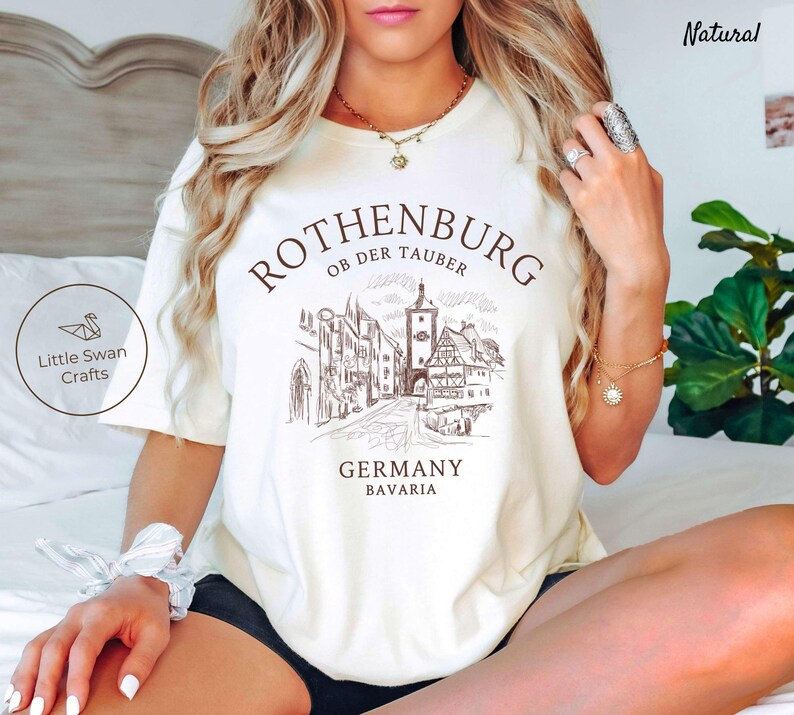 Pode incluir: Camiseta branca com um gr&aacute;fico marrom da cidade de Rothenburg ob der Tauber na Alemanha. O texto "Rothenburg ob der Tauber" est&aacute; acima do gr&aacute;fico, e o texto "Alemanha Baviera" est&aacute; abaixo do gr&aacute;fico.