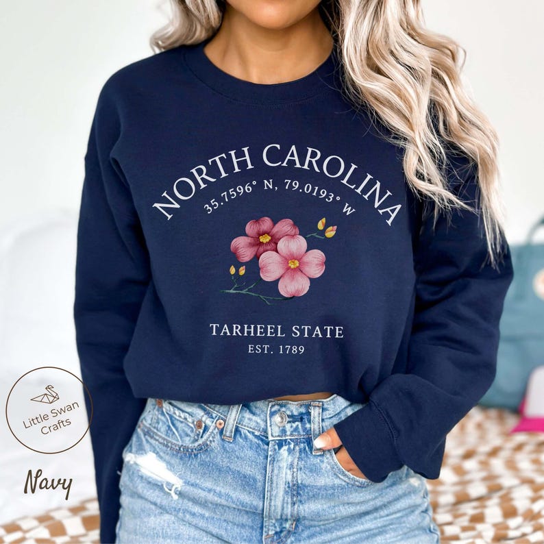 May include: Navy blue sweatshirt featuring the text "NORTH CAROLINA" with coordinates and a floral design. Below, it reads "TARHEEL STATE EST. 1789". The sweatshirt also has the "Little Swan Crafts" logo and the word "Navy".
