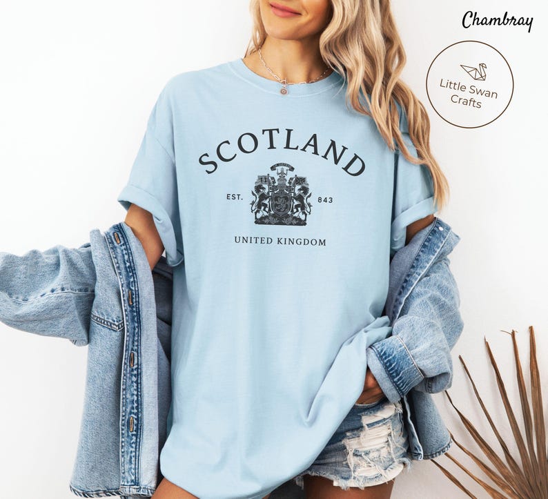 Pode incluir: Camiseta azul clara com a palavra "SCOTLAND" em arco e um desenho de bras&atilde;o abaixo. O texto "EST. 843 UNITED KINGDOM" est&aacute; abaixo do bras&atilde;o. Parte de uma cole&ccedil;&atilde;o de roupas.