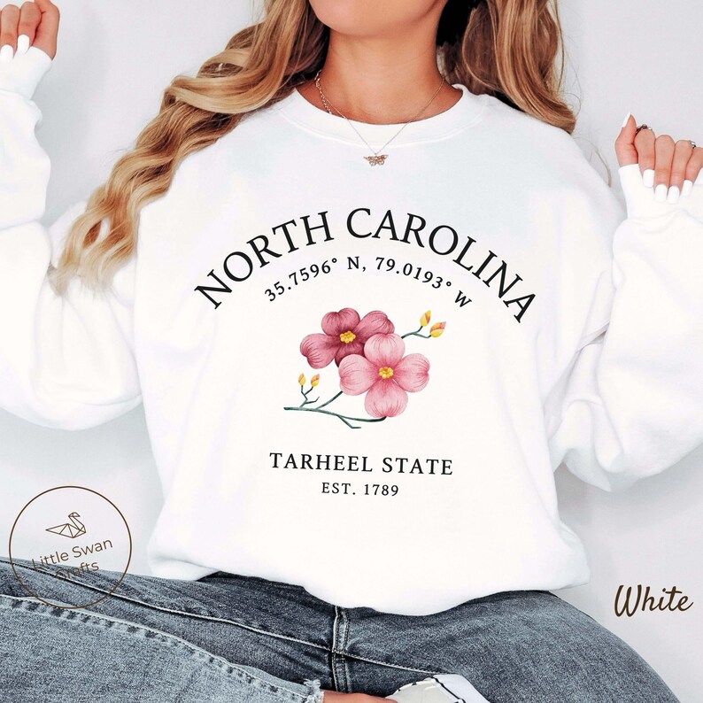 May include: White sweatshirt with the text "NORTH CAROLINA" and coordinates. Below the text is a floral design with pink flowers. The words "TARHEEL STATE" and "EST. 1789" are also printed on the sweatshirt. The word "White" is printed on the bottom right.