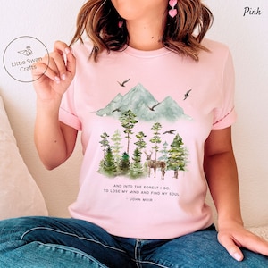 Pode incluir: Camiseta rosa com uma ilustra&ccedil;&atilde;o em aquarela de uma cordilheira, &aacute;rvores e um veado. O texto "AND INTO THE FOREST I GO TO LOSE MY MIND AND FIND MY SOUL - JOHN MUIR -" est&aacute; impresso abaixo da ilustra&ccedil;&atilde;o.