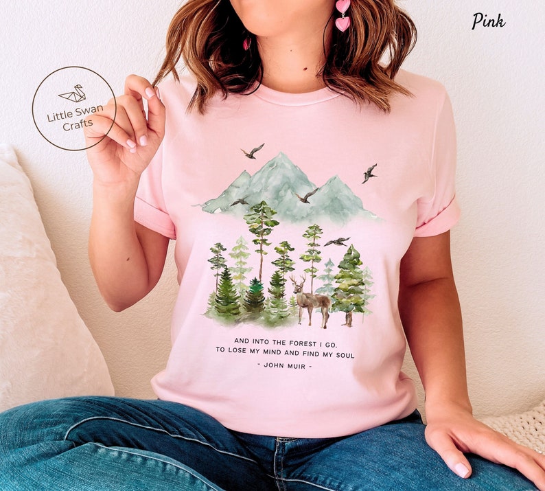 Pode incluir: Camiseta rosa com uma ilustra&ccedil;&atilde;o em aquarela de uma cordilheira, &aacute;rvores e um veado. O texto "AND INTO THE FOREST I GO TO LOSE MY MIND AND FIND MY SOUL - JOHN MUIR -" est&aacute; impresso abaixo da ilustra&ccedil;&atilde;o.