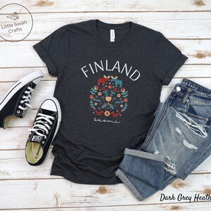 May include: Dark grey heather t-shirt with a white graphic of a floral design with the word "FINLAND" above it and the word "suomi" below it. The graphic features a moose, a fox, a bear, and a bird.