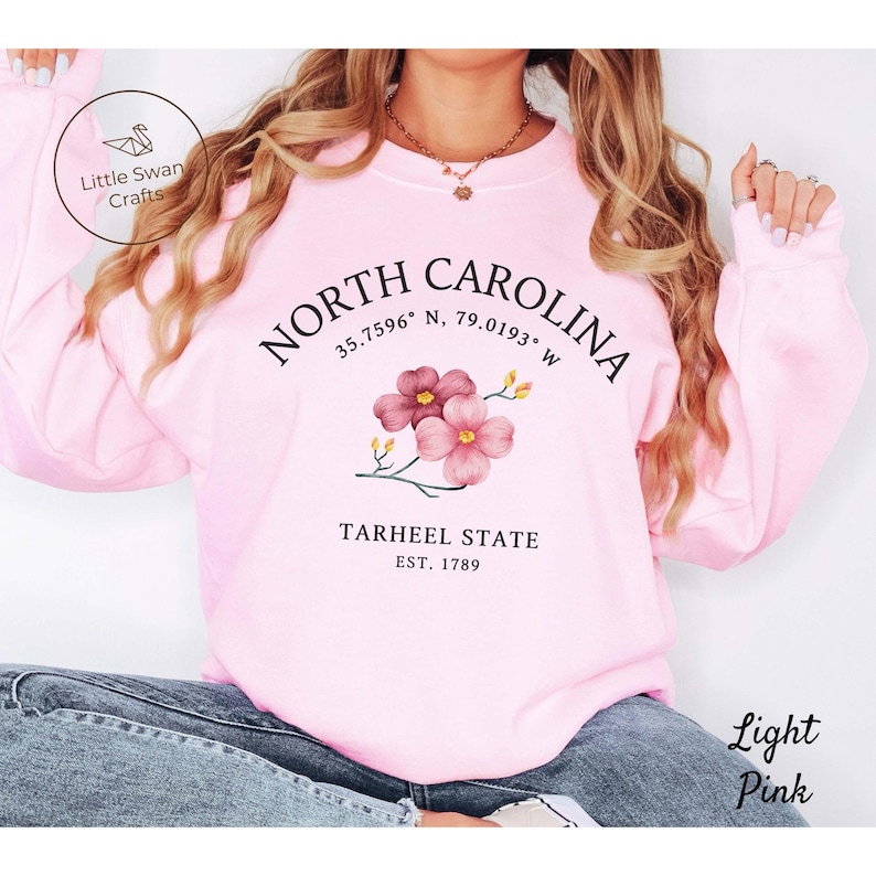 May include: Light pink sweatshirt with the text "NORTH CAROLINA" and coordinates, a floral design, and "TARHEEL STATE EST. 1789". The sweatshirt also has the text "Light Pink" on the bottom right.