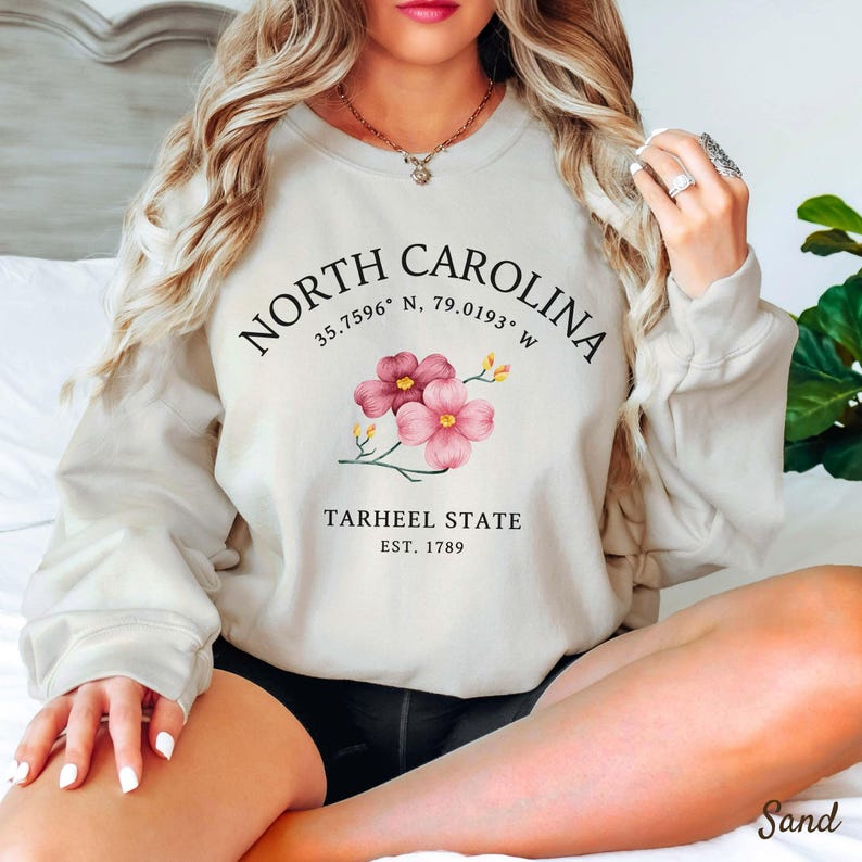May include: Sand-colored sweatshirt with the text "NORTH CAROLINA" arched across the top, coordinates, and "TARHEEL STATE EST. 1789" below. Features a floral design with pink flowers. The word "Sand" is at the bottom right.