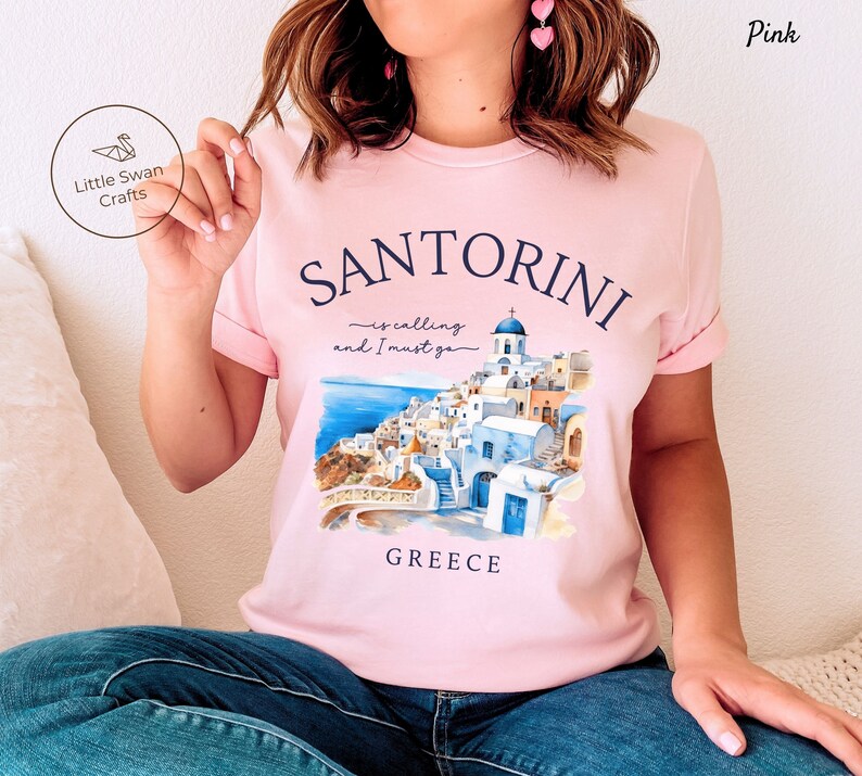 May include: Pink t-shirt with a watercolor illustration of Santorini, Greece. The text on the shirt reads "Santorini is calling and I must go".