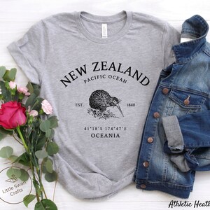 Peut inclure: T-shirt gris avec un graphique noir d'un kiwi et le texte "NEW ZEALAND PACIFIC OCEAN EST. 1840 41&deg;18&prime;S 174&deg;47&prime;E OCEANIA"