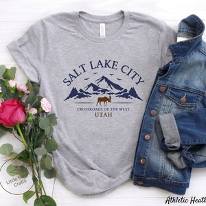May include: A gray t-shirt with a graphic design featuring a mountain range, a moose, and the text "Salt Lake City, Crossroads of the West, Utah".