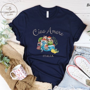 May include: A navy blue t-shirt with a watercolor illustration of a scooter, a house, and greenery. The text "Ciao Amore Italia" is printed on the shirt.