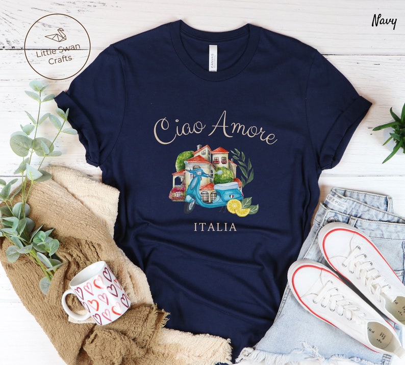May include: A navy blue t-shirt with a watercolor illustration of a scooter, a house, and greenery. The text "Ciao Amore Italia" is printed on the shirt.