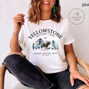 May include: White t-shirt with a Yellowstone National Park graphic. The design features a moose, mountains, and trees, with text indicating the park's name, location, and establishment year. The shirt also has the text "Little Swan Crafts".