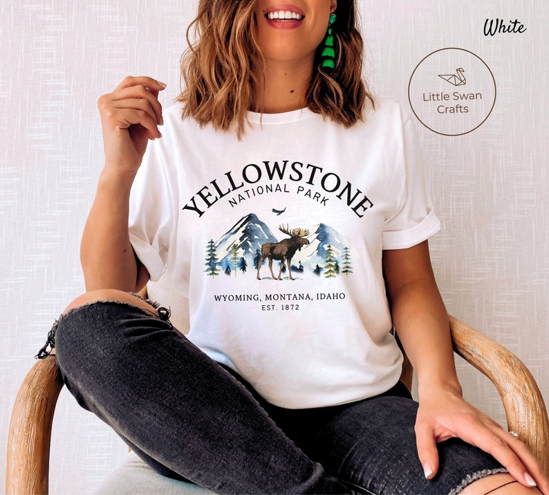 May include: White t-shirt with a Yellowstone National Park graphic. The design features a moose, mountains, and trees, with text indicating the park's name, location, and establishment year. The shirt also has the text "Little Swan Crafts".