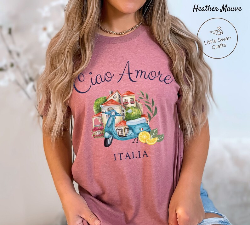May include: A heather mauve t-shirt with a graphic of a blue scooter in front of a small Italian village. The text "Ciao Amore" is above the scooter and "ITALIA" is below.