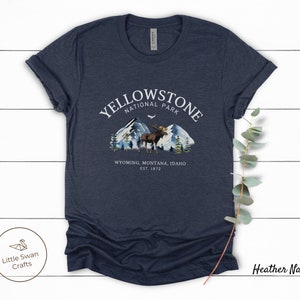 May include: Heather navy t-shirt with a graphic of Yellowstone National Park. The design features mountains, trees, and a moose, with the text "YELLOWSTONE NATIONAL PARK" and "WYOMING, MONTANA, IDAHO EST. 1872". The shirt has rolled-up sleeves.