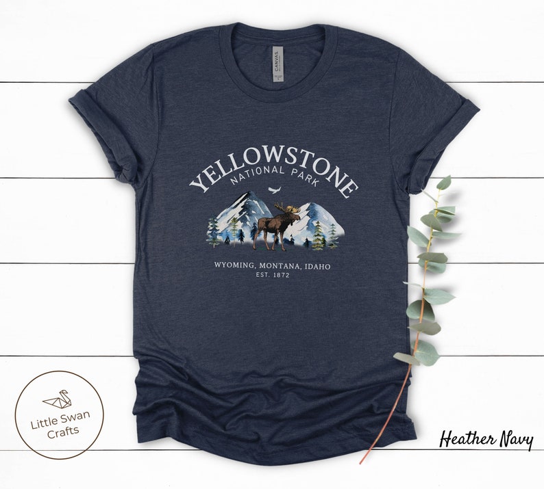 May include: Heather navy t-shirt with a graphic of Yellowstone National Park. The design features mountains, trees, and a moose, with the text "YELLOWSTONE NATIONAL PARK" and "WYOMING, MONTANA, IDAHO EST. 1872". The shirt has rolled-up sleeves.