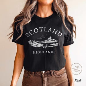May include: Black t-shirt featuring a white illustration of the Scottish Highlands, with a castle, sheep, and the text "SCOTLAND HIGHLANDS". The shirt is made of a soft fabric.