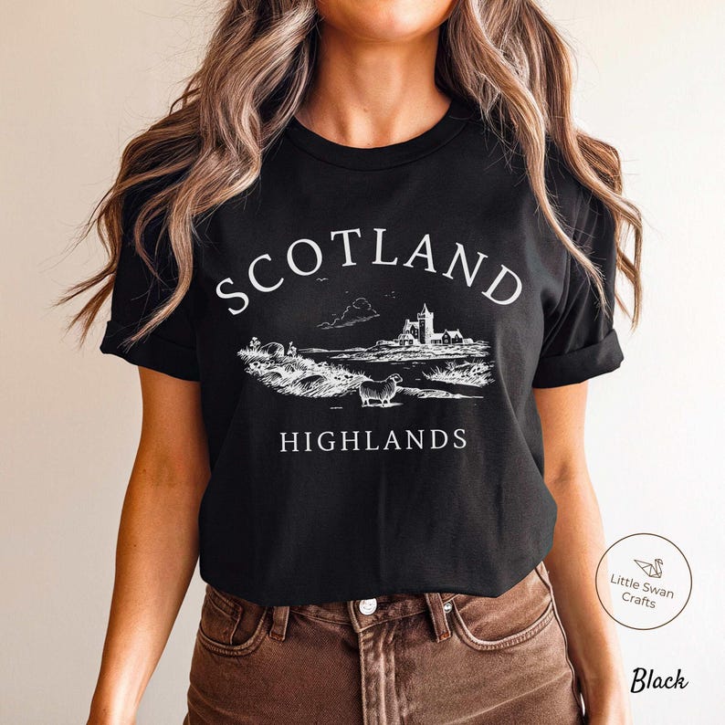 May include: Black t-shirt featuring a white illustration of the Scottish Highlands, with a castle, sheep, and the text "SCOTLAND HIGHLANDS". The shirt is made of a soft fabric.