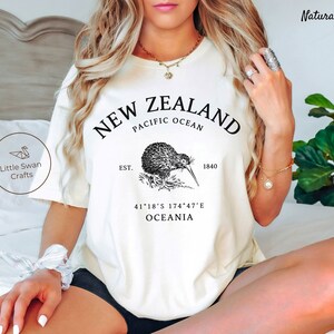 Peut inclure: T-shirt blanc avec un motif graphique noir repr&eacute;sentant un kiwi, le texte "NEW ZEALAND", "PACIFIC OCEAN", "EST. 1840", "41&deg;18'S 174&deg;47'E", et "OCEANIA".