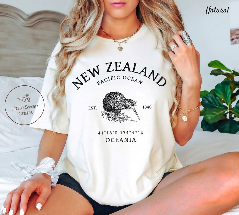 Peut inclure: T-shirt blanc avec un motif graphique noir repr&eacute;sentant un kiwi, le texte "NEW ZEALAND", "PACIFIC OCEAN", "EST. 1840", "41&deg;18'S 174&deg;47'E", et "OCEANIA".