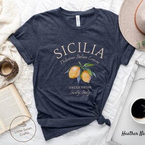 Peut inclure: Un t-shirt bleu marine chin&eacute; avec un motif graphique repr&eacute;sentant le texte "SICILIA Delicious Italian Lemons" et une illustration aquarelle de citrons sur une branche. Le texte "FRESH FROM Sicily, Italy" est &eacute;galement inclus dans le design.