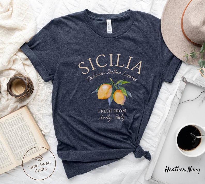 Peut inclure: Un t-shirt bleu marine chin&eacute; avec un motif graphique repr&eacute;sentant le texte "SICILIA Delicious Italian Lemons" et une illustration aquarelle de citrons sur une branche. Le texte "FRESH FROM Sicily, Italy" est &eacute;galement inclus dans le design.
