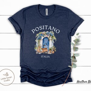 Puede incluir: Camiseta azul marino jaspeada con una ilustraci&oacute;n de acuarela de una puerta azul con flores y un scooter delante. El texto "Positano" e "Italia" est&aacute; impreso en la camiseta.