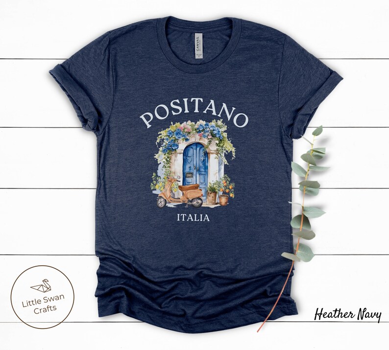 Puede incluir: Camiseta azul marino jaspeada con una ilustraci&oacute;n de acuarela de una puerta azul con flores y un scooter delante. El texto "Positano" e "Italia" est&aacute; impreso en la camiseta.