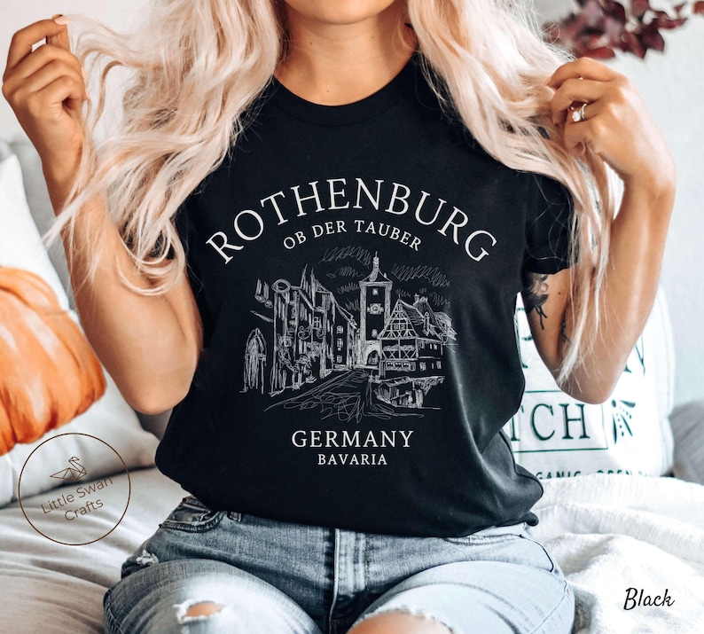 Pode incluir: Camiseta preta com um gr&aacute;fico branco da cidade de Rothenburg ob der Tauber, Alemanha. O gr&aacute;fico inclui um esbo&ccedil;o dos edif&iacute;cios e ruas da cidade. O texto "Rothenburg ob der Tauber" est&aacute; escrito em branco, com uma fonte cursiva. O texto "Alemanha Baviera" est&aacute; escrito em branco, com uma fonte sem serifa.