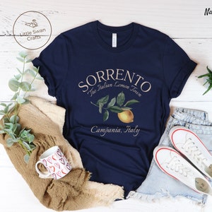 May include: Navy blue t-shirt with the text "Sorrento The Italian Lemon Town Campania, Italy" and a lemon illustration. The shirt is displayed with white sneakers and a mug with red hearts.