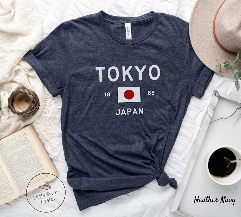 K&ouml;nnte beinhalten: Heather-Marineblaues T-Shirt mit wei&szlig;em Text, der "TOKYO" und "JAPAN" mit der japanischen Flagge dazwischen lautet. Die Zahlen 18 und 68 befinden sich auf beiden Seiten der Flagge.